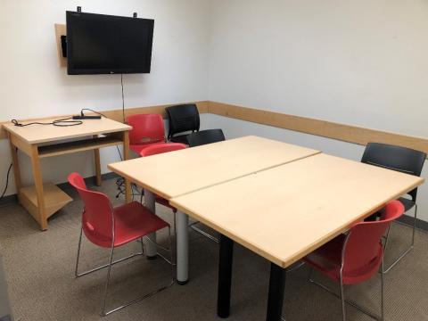 Group study room with two tables pushed together. Some chairs are stacked in a corner next to a second table to hold laptops to connect to a monitor on the wall. Three chairs are around the main table. 