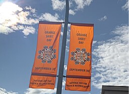 Two orange banners are attached to pole reaching into the blue sky and the sun behind them. Both banners read Orange Shirt Day, September 30. National Day for Truth and Reconciliation. An image on each banner also reads, “Every Child Matters”