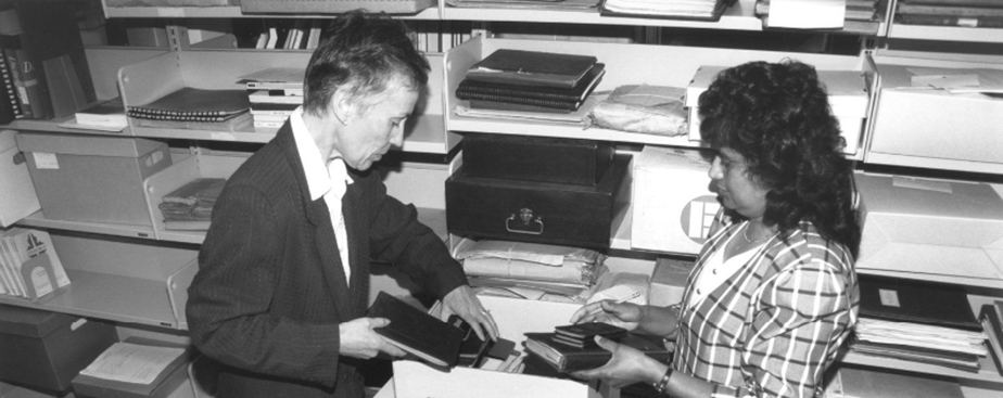 Joan Mitchell and Doreen Udit putting books in a box in front of a shelving unit.