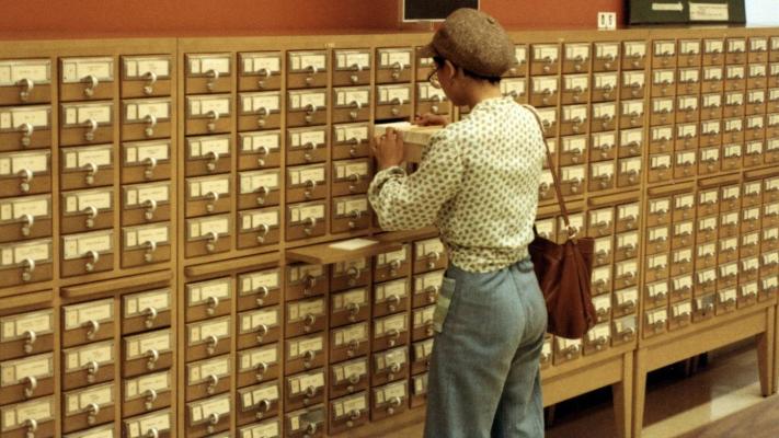 Woman standing in front of library card catalogue looking through the cards in an open drawer.