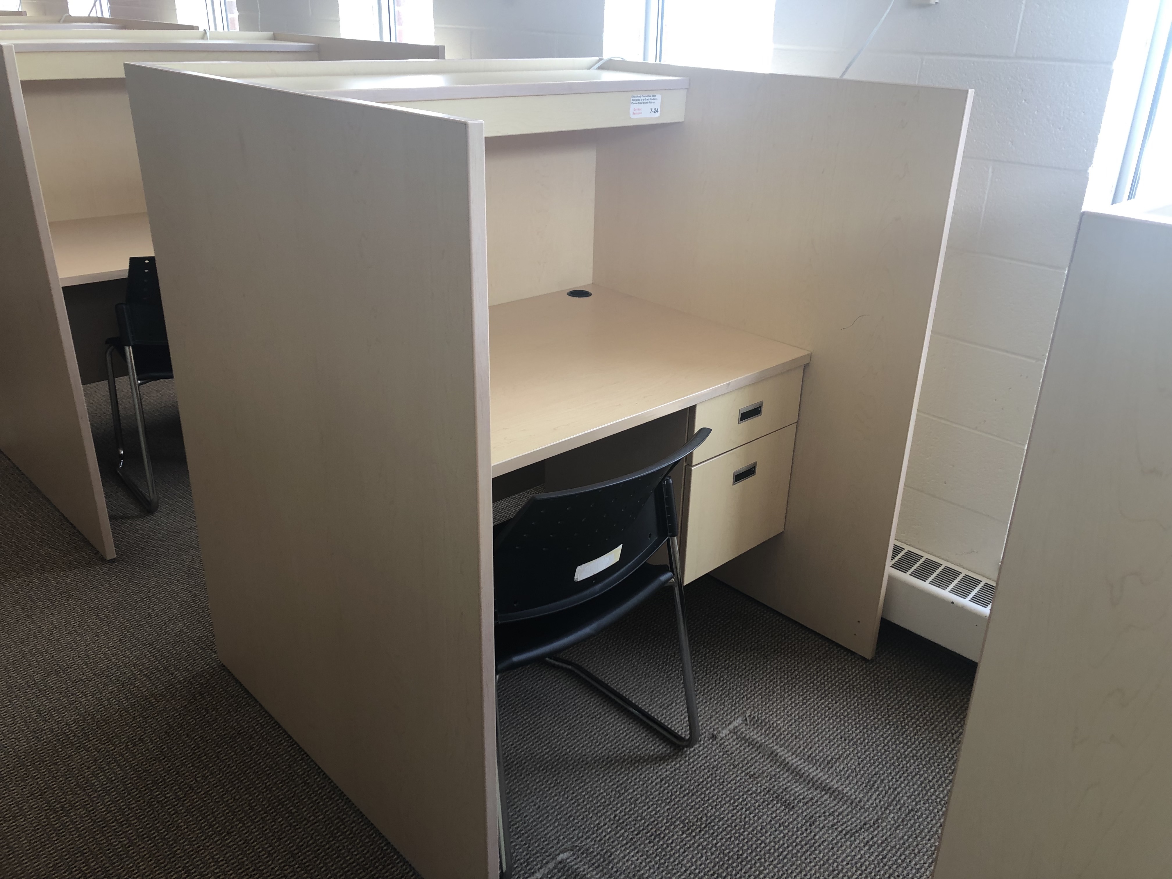 Image of grad carrel. On upper floors, along an outer wall, near windows. Desk with side panels on either side for privacy. A lockable desk drawer. Not much space under desk. 
