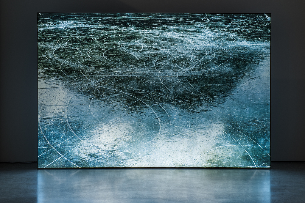 This image is an installation view of Laura Millard’s lightbox entitled Lac des Arcs. The image on the lightbox is that of a frozen pond on which bodies have skated. White blade markings depict circular and curved lines suggesting the aftermath of a hockey game or figure skating event. The top colours in the image are darker while the bottom comprises varying shades of light blues. Displayed in an unlit space, the lightbox casts a glow and mirrored reflection of the piece on the floor
