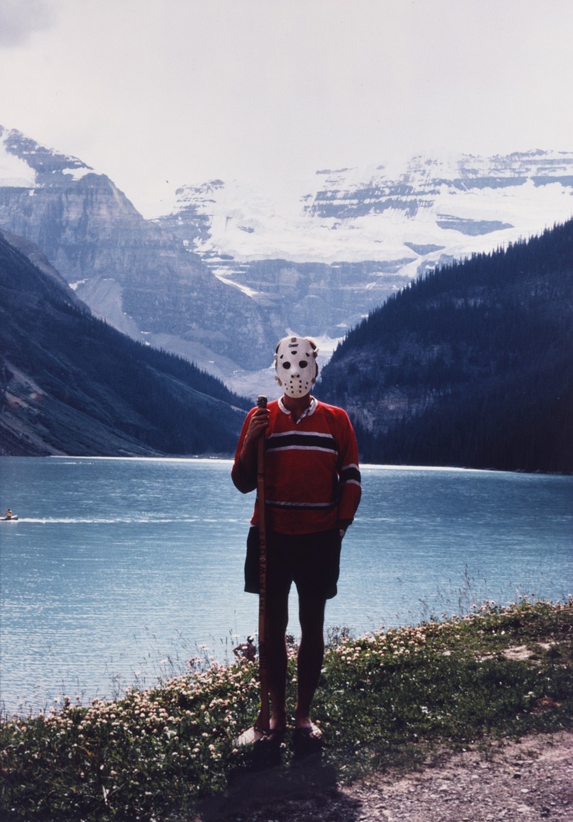 Photograph of a man in a red jersey holding a hockey stick, whose face is completely covered by a traditional white goalie mask with holes for the eyes. The man is standing in front of Lake Louise, a turquoise glacial lake, with snow covered mountains in the background.
