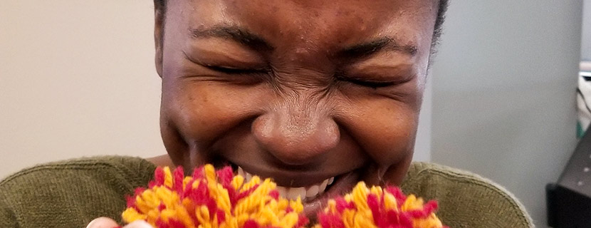 The delight of making. A student with dark skin on rising dimpled cheeks, a crinkled nose, and eyes closed tightly, smiles with delight while holding pompoms of red and gold strands of yarn, made at the Library Maker space. Music accompaniment is at the next button. A text description of audio follows at the button labelled Click for Audio alternative