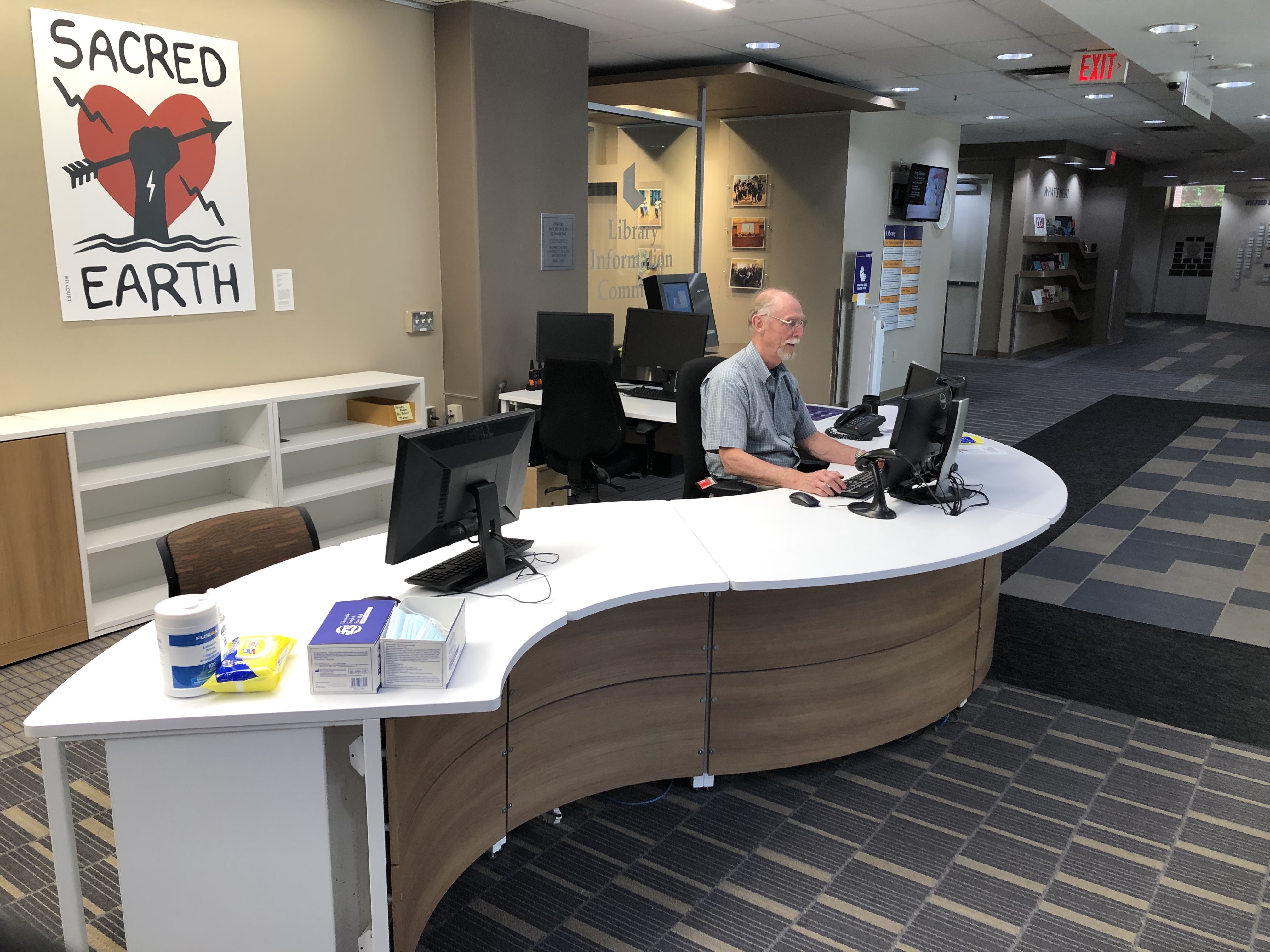 The user services desk, staffed with a library employee. The desk is large and curvy. On the wall is art “Sacred Earth” with hand holding arrow over a heart.