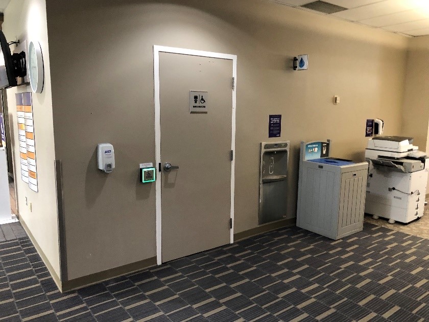 The entrance to an individual use washroom. It has a lever handle and a large square button for automatically opening the door. It glows green if open or red if the door is locked. It is close to a water bottle filling station and photocopiers and printing stations