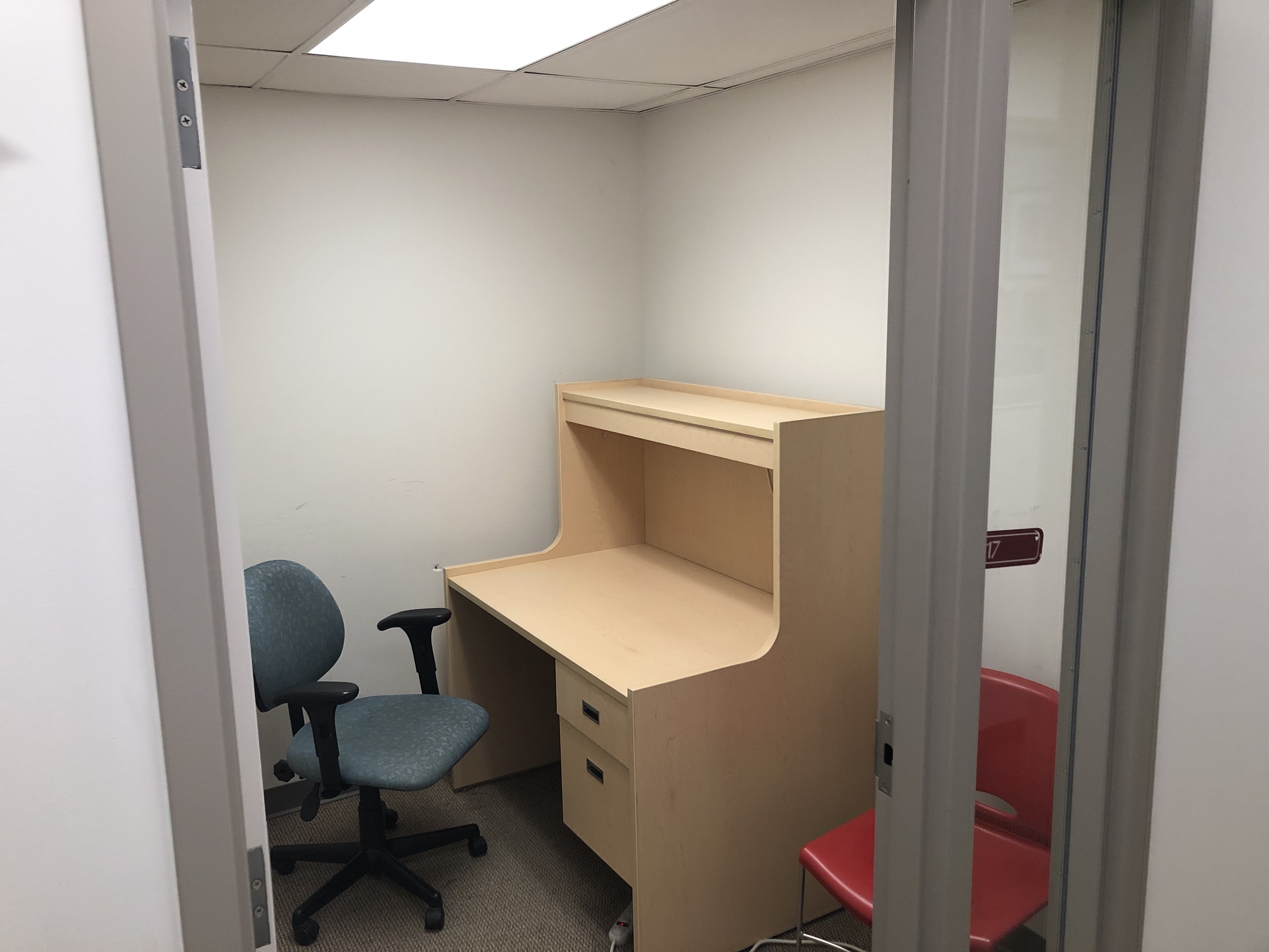 A private study room with a chair and desk.  The desk has draws. There is a tall, narrow window next to door