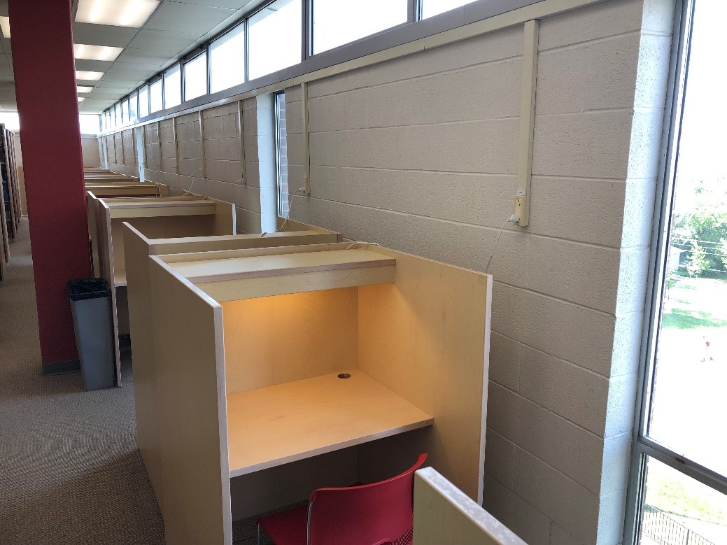 A line of study carrels along the outer wall of the library. There are partitions on the left and right side of these desks, however, these desks are arranged face front to back with other desk. They are still separated by a partition for privacy.