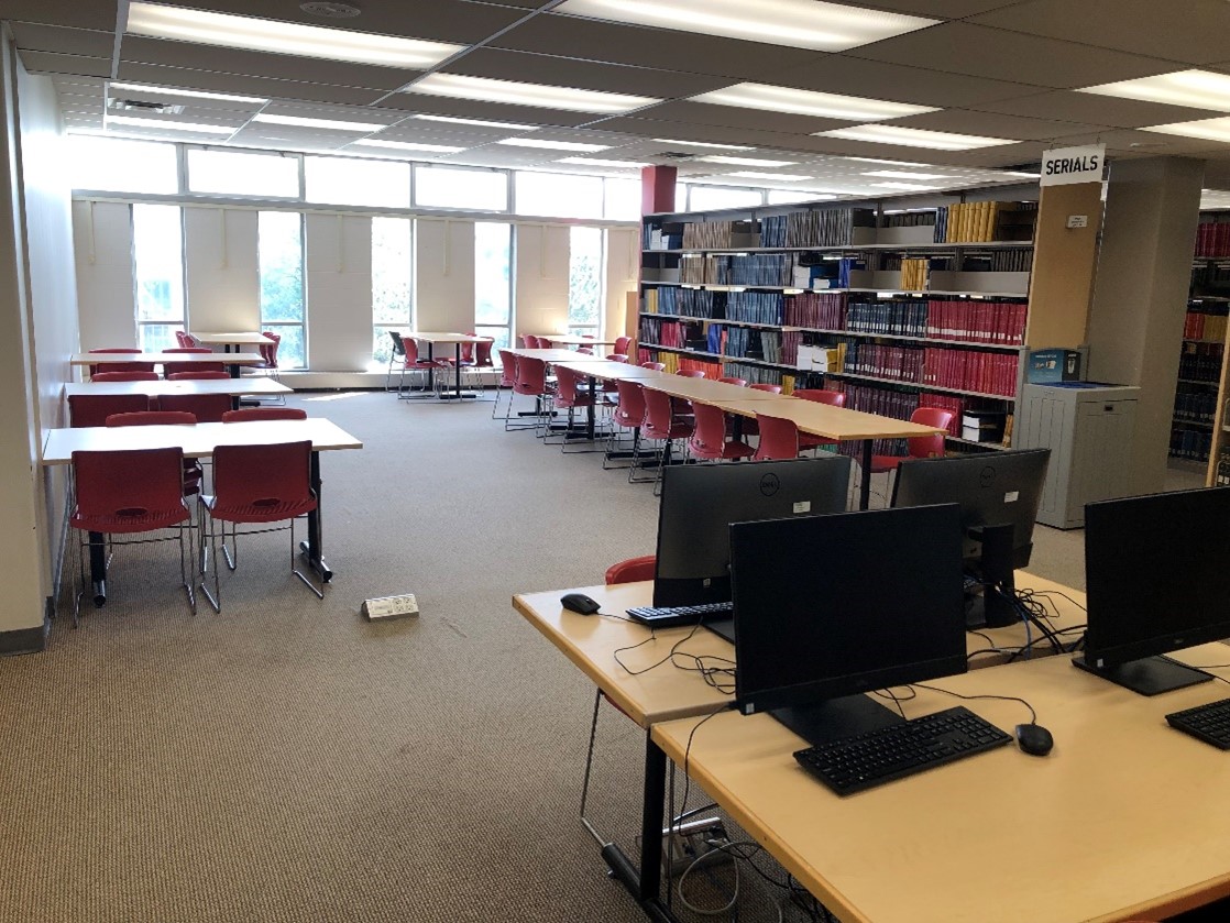 An area on the 4th floor. A cluster of workstations on desks. Nearby are several tables with four chairs around each. There’s also a long desk with chairs on either side. Large bookshelves are close that hold print journals. On the ground is an electrical outlet awkwardly sticking out of the floor. It would be at tripping hazard.