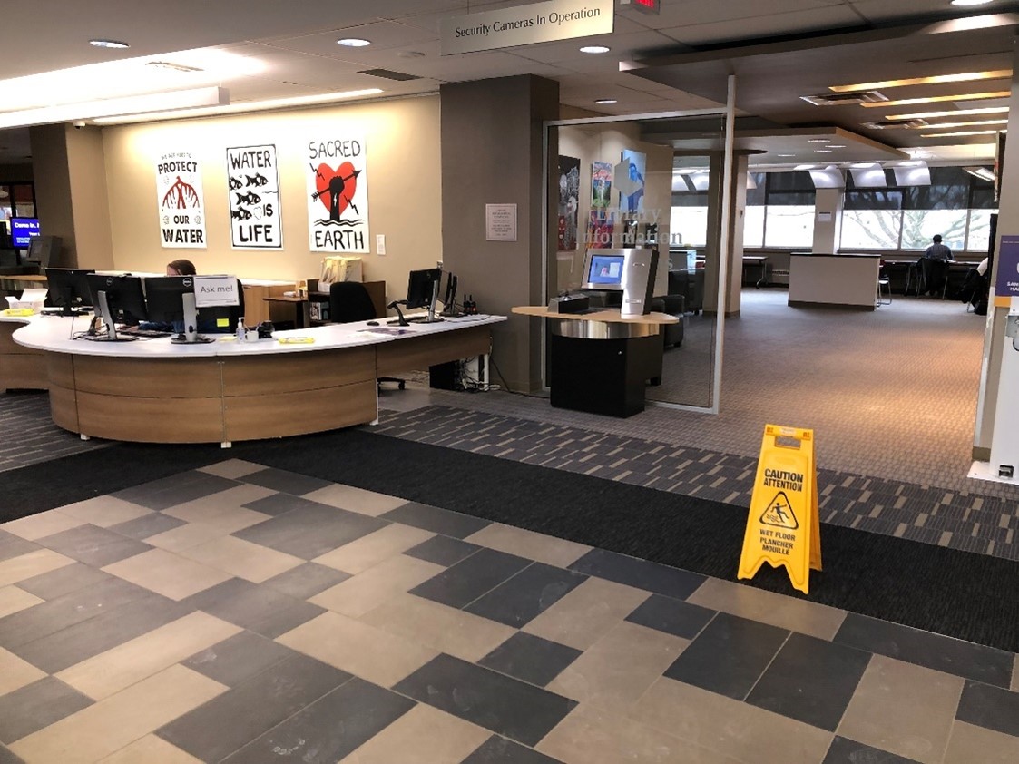 Lobby of the library at 10 o’clock is the large user services desk. Through 12 o’clock is large room with a variety of seating and desks. On the wall is art. Some says ‘Water is life’. A signs says ‘Security cameras in operation’
