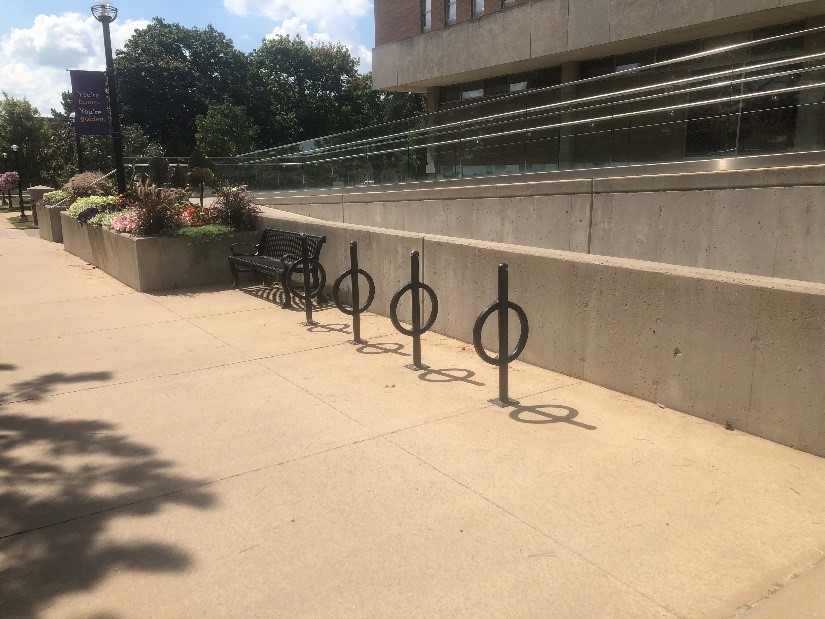 Metal bench, and four bike racks. Each rack is a separate metal post with rounded bar. In this picture there are no bikes. Behind them is a concrete barrier that runs along side of the ramp for entering the library.
