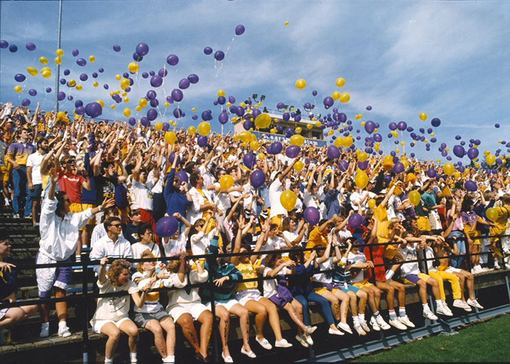 Crowd sitting on bleachers throwing yellow and purple balloons in the air.