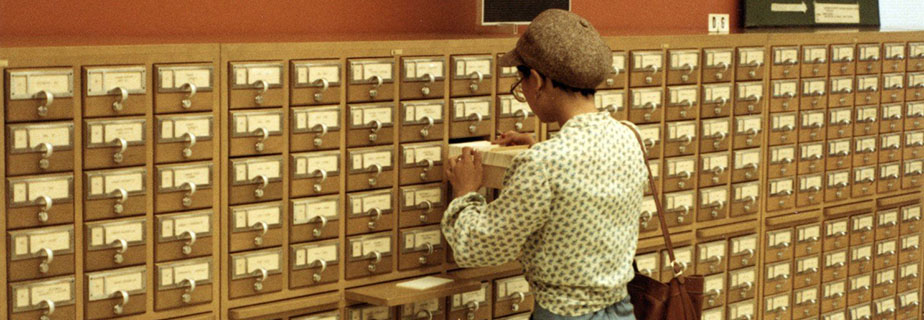Woman standing in front of library card catalogue looking through the cards in an open drawer.