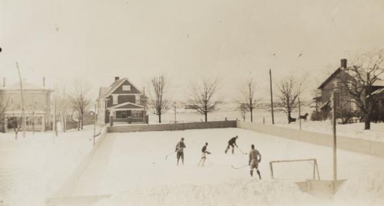 Four people playing hockey on an ice rink at the Evangelical Lutheran Seminary of Canada. Albert Street can be seen in the background with fields beyond.