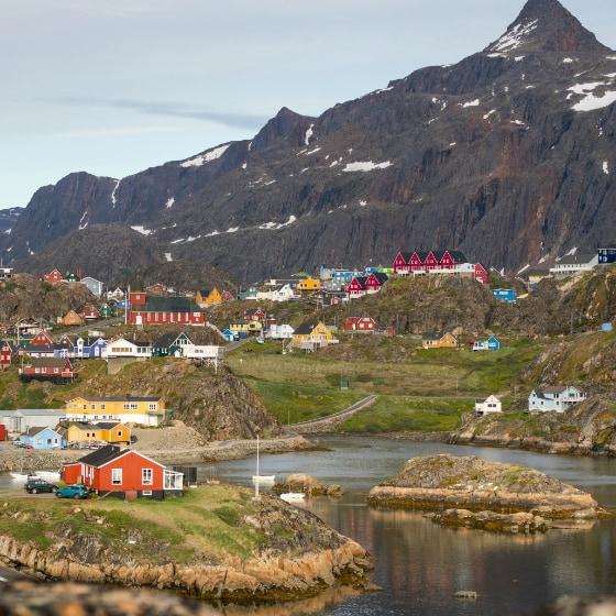 A view of Greenland, showing a snow-covered mountain in the background, colourful houses on hills in the middle zone, and an inlet of water in the foreground.
