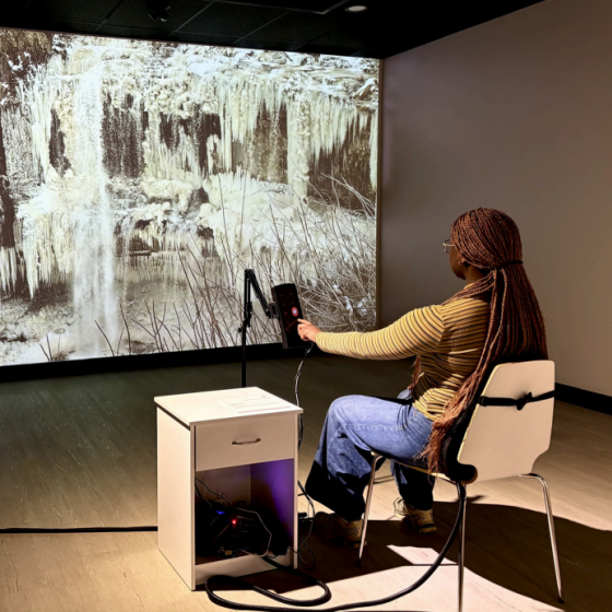 A person in library art gallery sits in a chair in front of a large image of a frozen waterfall. The chair has a vibrating back.