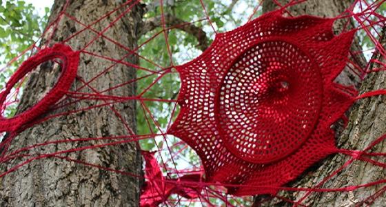 Various circular shapes knitted from red acrylic yarn are attached to the body of a large tree with red string.