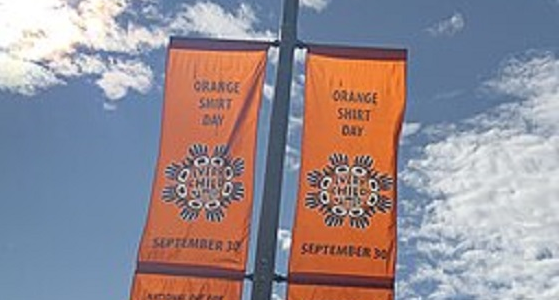 Two orange banners are attached to a lamppost with blue sky and the sun behind them. Both banners read Orange Shirt Day, September 30. National Day for Truth and Reconciliation. An image on each banner also reads, “Every Child Matters” 