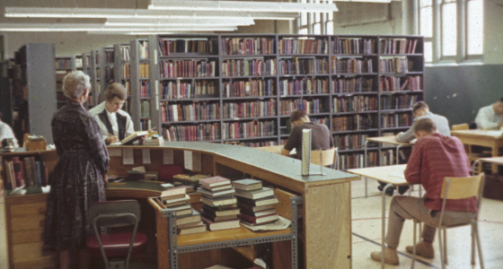 Staff stand at the circulation desk in the Waterloo College Library while students work at individual desks nearby. Rows of bookshelves can be seen in the background.