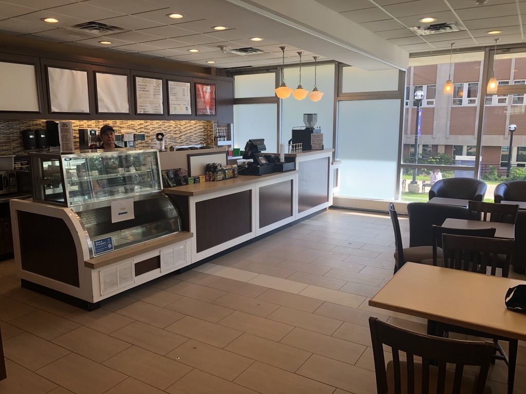 Coffee shop when it is open. There is a sandwich board with hours. The floor of the coffee shop is titled while the lobby is carpeted. There is a counter to order and usually two employees. There are chairs and tables, and also some comfy chairs. Space is enclosed by windows.