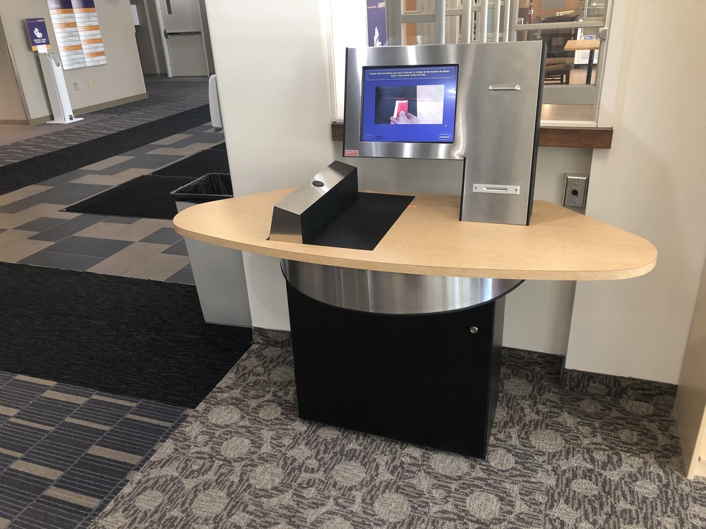 A self-service library kiosk.  The kiosk features a large touch-screen interface mounted on a silver panel. It is placed on a light wooden desk. The screen displays a webpage with instructions. It is on a carpeted surface. Items can be checked out manually at the user services desk.