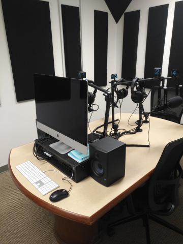 Podcast studio desk with Mac workstation, speakers, and microphones. Sound bafflers on the wall